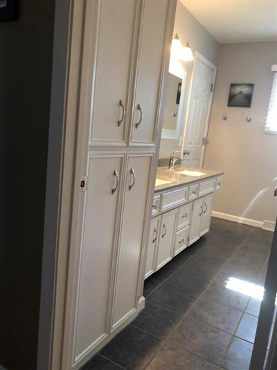 Bathroom with white cabinetry, a double-sink vanity, gray countertop, and dark tiled floor, featuring a well-lit mirror and ample storage space.