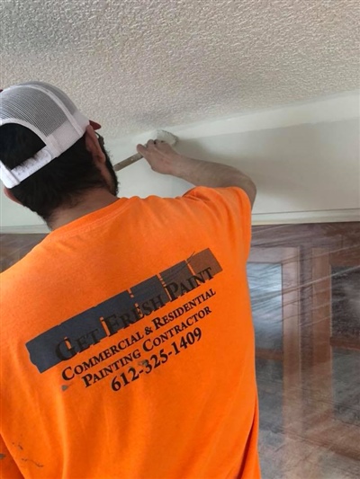 A worker in an orange shirt with company information is painting a ceiling.