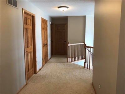 A carpeted hallway with light beige walls, wooden doors, and a wooden railing leading to a staircase.