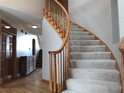 A carpeted spiral staircase with wooden railings, leading up from a hallway with wooden flooring and white walls.