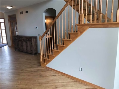 A wooden staircase with railings leading up from a hallway with wooden flooring, white walls, and a wooden cabinet against the wall.